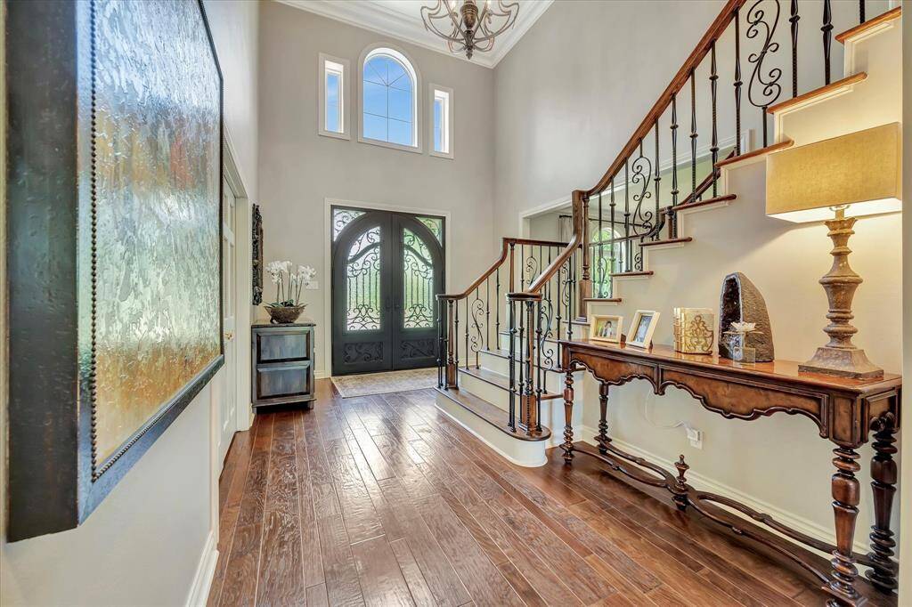 This elegant entryway features a grand staircase with ornate iron railings, hardwood floors, and a high ceiling. A large decorative front door and ample natural light create a welcoming atmosphere. A stylish console table and art adorn the space, adding warmth and sophistication.