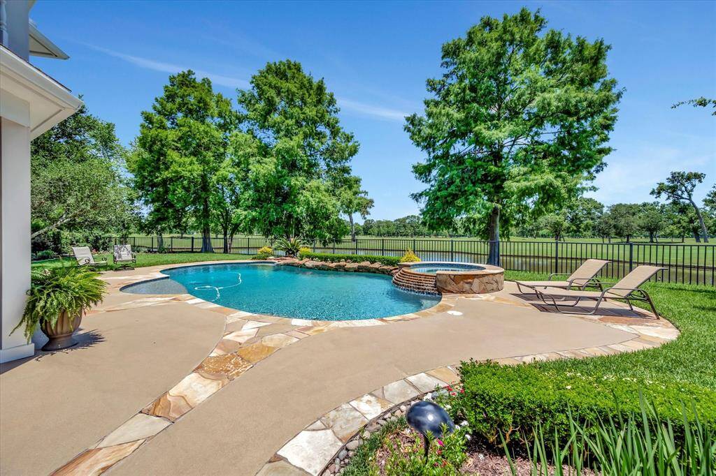 This beautiful property features a spacious backyard with a well-maintained lawn, a relaxing pool with a stone border, and a scenic view of a pond. The covered patio offers ample space for outdoor seating, perfect for enjoying the serene surroundings.