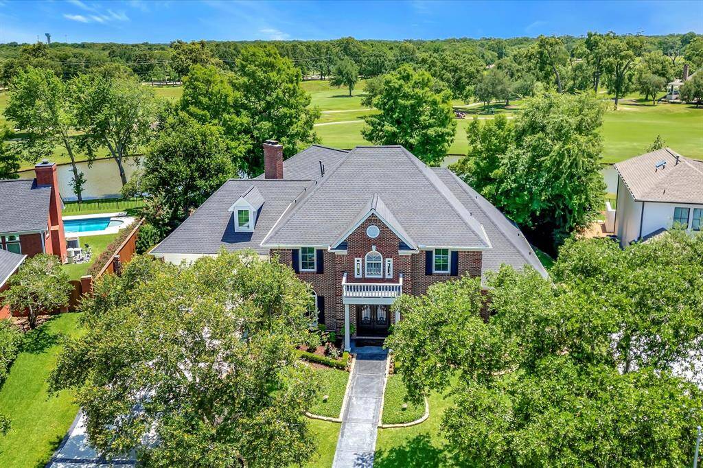 This photo showcases a spacious, elegant brick home with a well-manicured lawn and mature trees. It features a large, private backyard and borders a lush green golf course, providing scenic views , cool breezes and privacy. Outdoor security cameras for peace of mind.