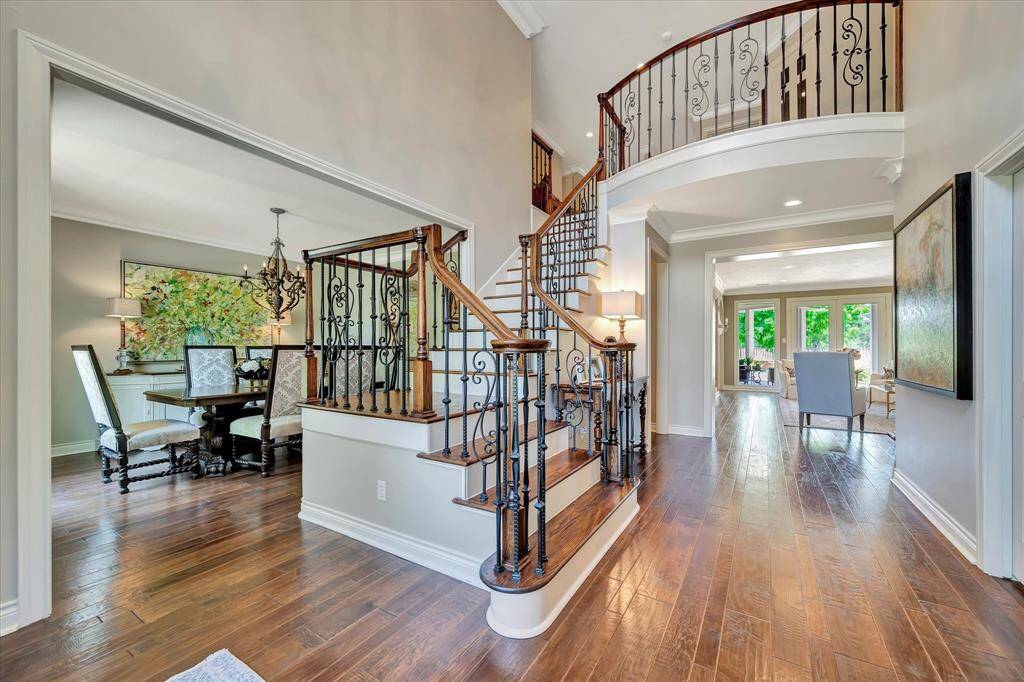 This inviting entryway features elegant hardwood floors and a grand staircase with ornate iron railings. The open layout flows into a formal dining room and a bright living area with large upgraded windows overlooking the backyard, creating a warm and welcoming atmosphere.