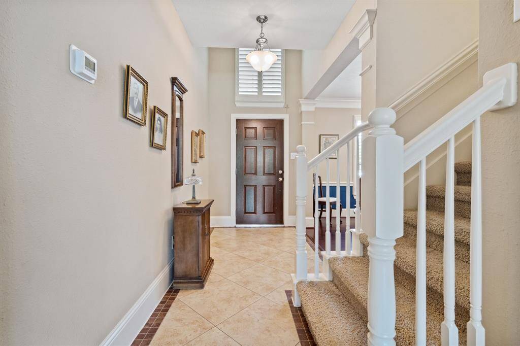 This inviting entryway features a warm, neutral color palette with ceramic tile flooring and a carpeted staircase. The space is well-lit with a stylish pendant light, and decorative wall frames add a personal touch. The wooden front door adds a classic element to the home.