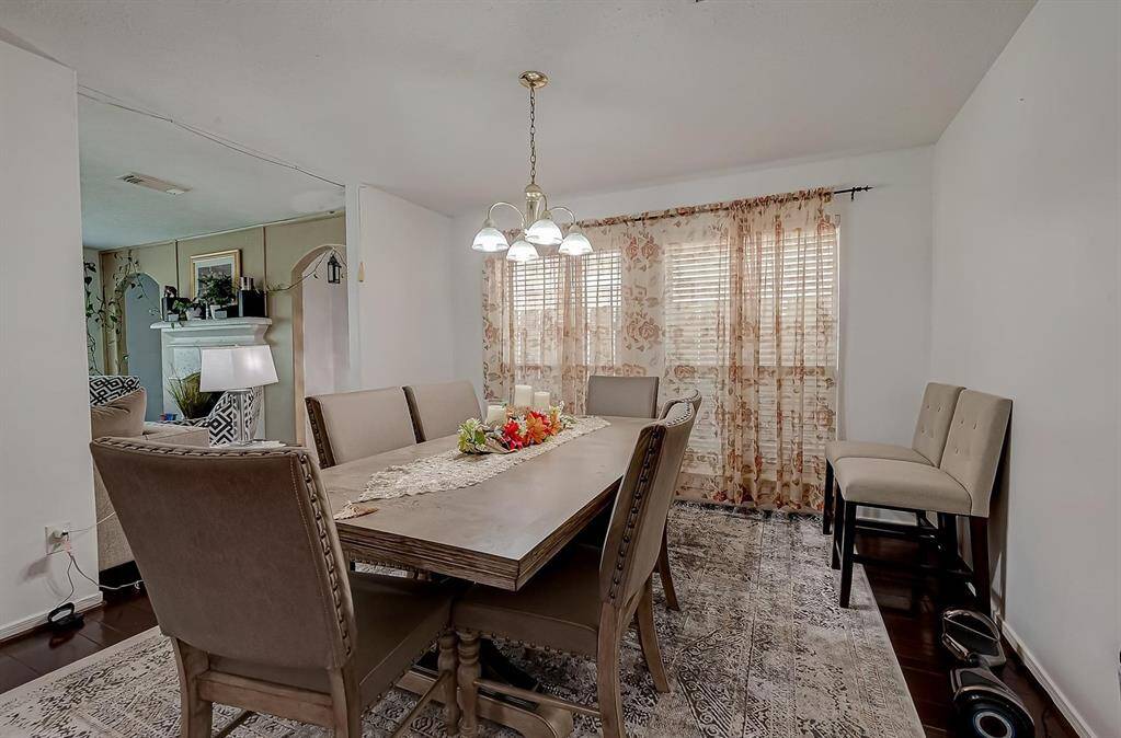 This photo shows a cozy dining area with a wooden table and upholstered chairs, illuminated by a stylish chandelier. The space has a warm, inviting ambiance with floral curtains and an adjacent living room featuring a fireplace.