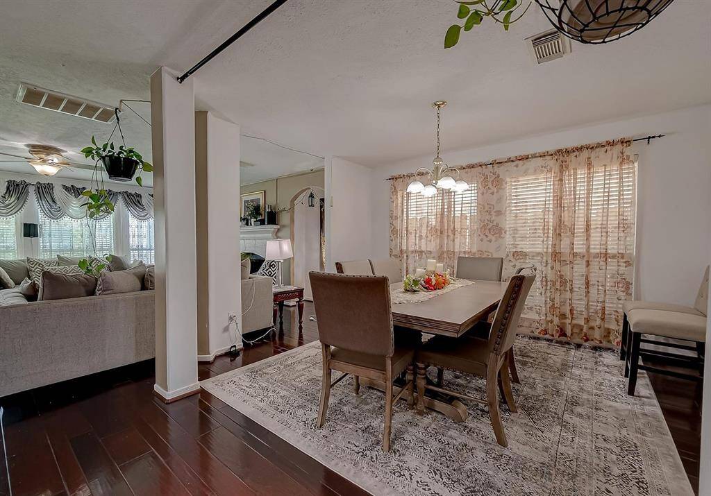 This photo showcases a cozy dining area with a wooden table and chairs set on a patterned rug. To the left, there's a glimpse of a comfortable living space with a sectional sofa. The room features hardwood floors, floral curtains, and hanging plants, creating a warm and inviting atmosphere.