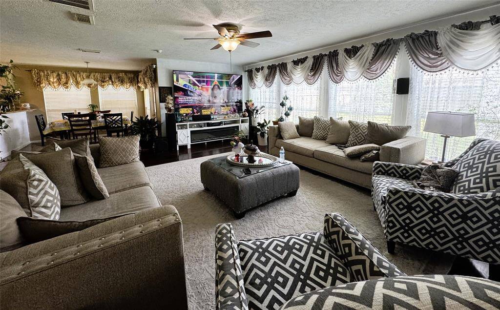 This spacious living room features large windows with elegant drapes, allowing plenty of natural light. It includes comfortable seating with plush sofas and patterned chairs, a ceiling fan, and a large TV. The open layout flows into the dining area, creating an inviting space for relaxation and entertainment.