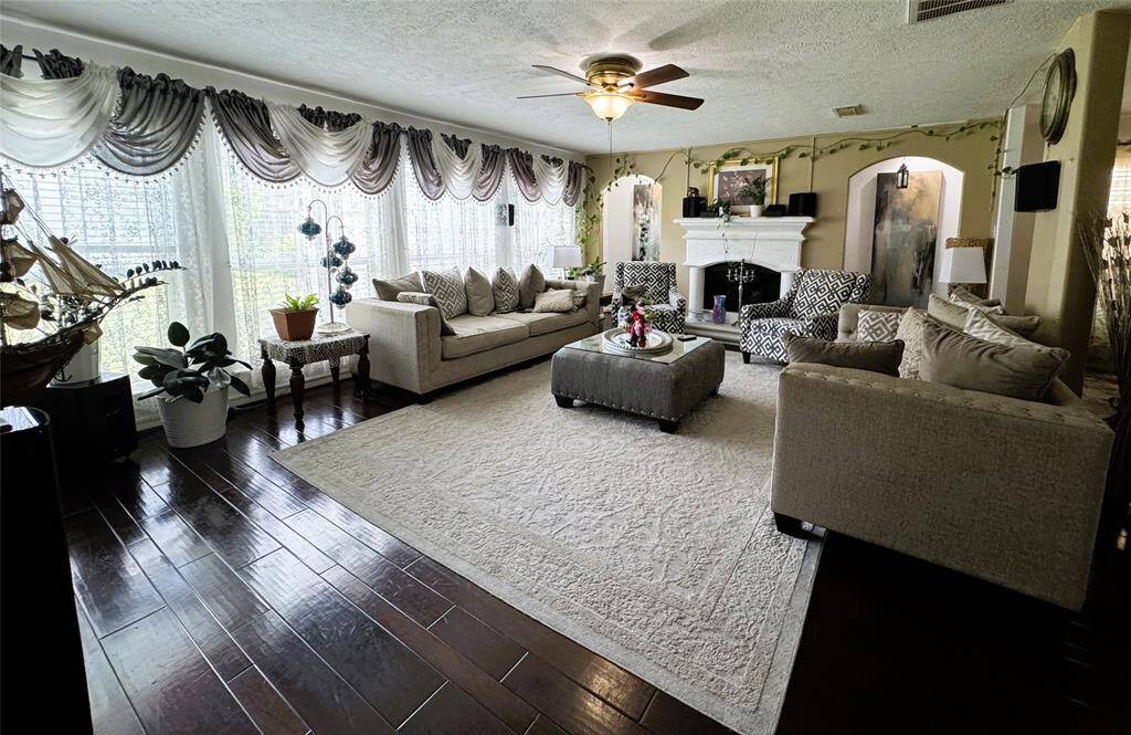 This spacious living room features elegant decor with large windows, plush seating, and a cozy fireplace. The hardwood floors and stylish ceiling fan add warmth, while the neutral colors and tasteful accents create a welcoming atmosphere.