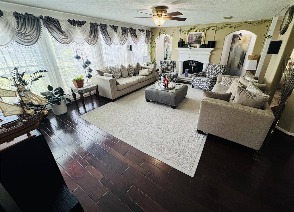 This spacious living room features elegant decor with plush sofas and a central coffee table on a light area rug. Large windows with ornate drapes provide ample light, while the dark wood flooring adds warmth. A ceiling fan and fireplace enhance comfort and ambiance.