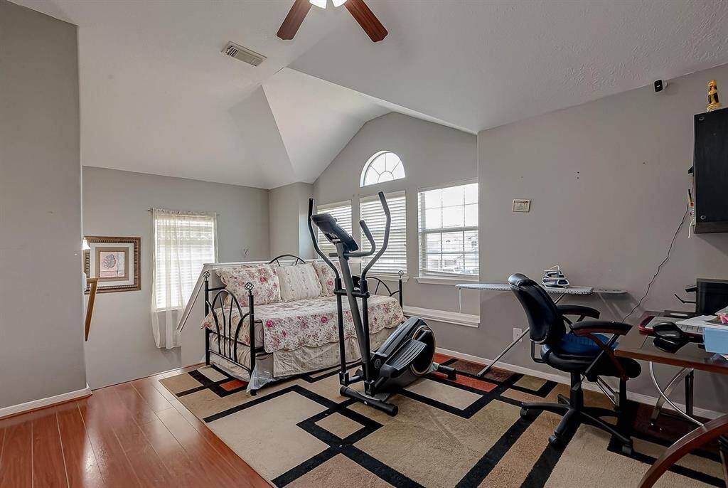 This room features a vaulted ceiling and ample natural light from large windows. It includes a bed with a floral bedspread, exercise equipment, a desk with a chair, and a modern rug on hardwood flooring, making it versatile for both relaxation and work.