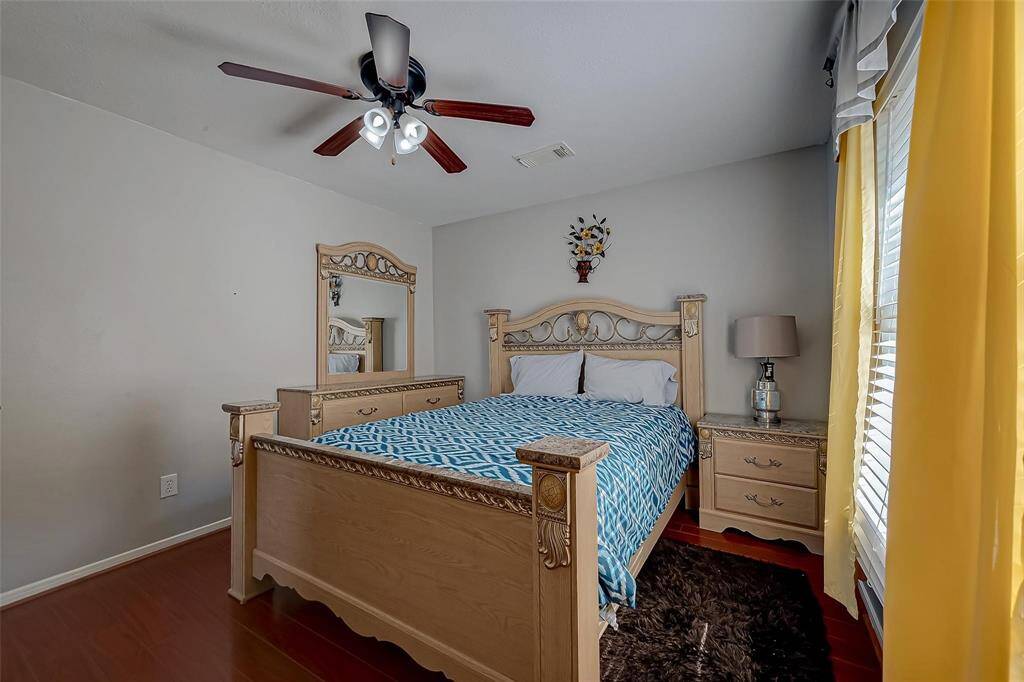 This cozy bedroom features a decorative wooden bed frame, matching nightstands, and a ceiling fan. The room is brightened by large windows with yellow curtains, creating a welcoming atmosphere.
