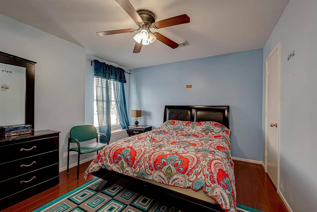 This cozy bedroom features a ceiling fan, hardwood floors, and a large window with blue curtains. The room is painted in a light blue tone and includes a double bed with a colorful quilt, a dresser, and a bedside table with a lamp.