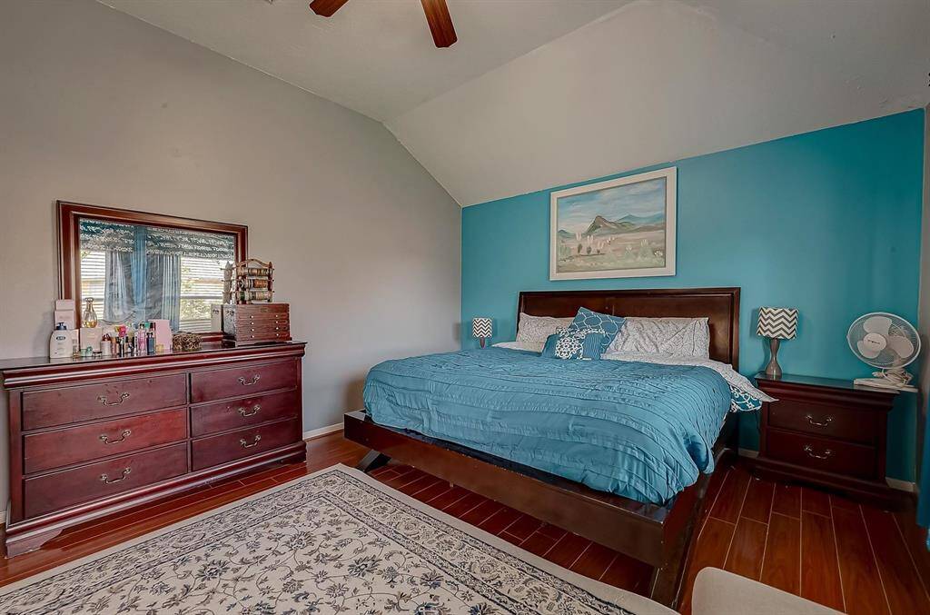 This bedroom features a cozy setup with a large bed, accented by a bold blue accent wall. It includes wooden furniture, such as a dresser and nightstands, offering ample storage. A patterned area rug complements the hardwood floors, creating a warm atmosphere.
