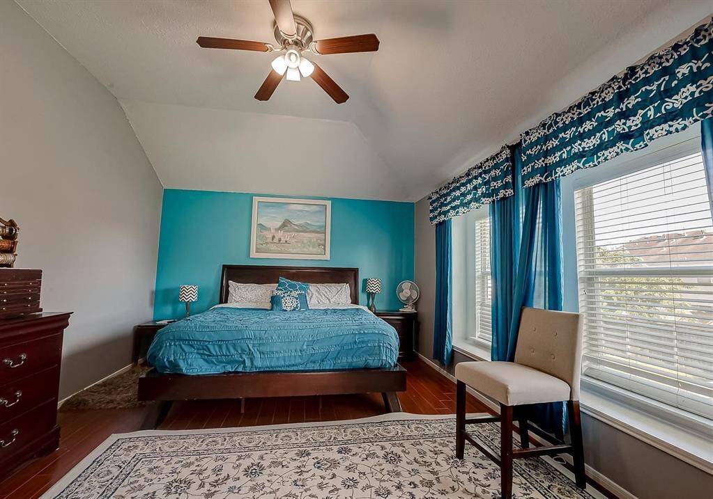 This bedroom features a vaulted ceiling with a ceiling fan, hardwood floors, and a bright accent wall. Large windows with elegant blue curtains provide ample natural light. The space includes a cozy bed, stylish decor, and a comfortable seating area.