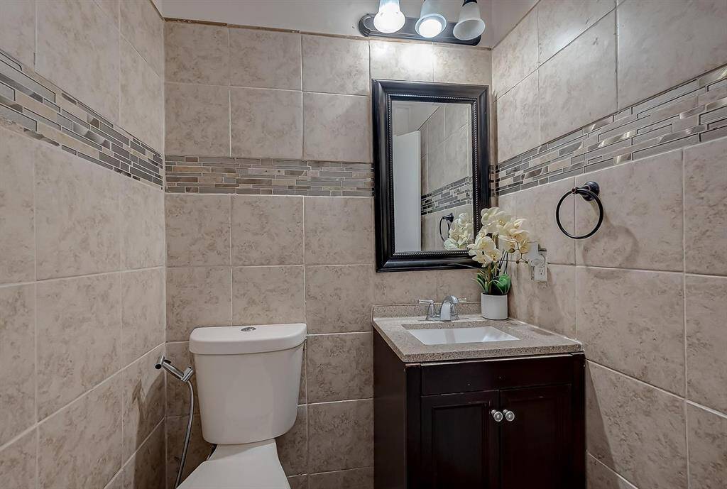 This bathroom features modern tile walls with a decorative mosaic stripe. It includes a sleek vanity with a dark wood finish, a granite countertop, and a mounted mirror. The lighting is contemporary, and there's a touch of elegance with a floral arrangement.