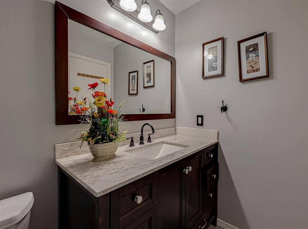 This bathroom features a modern vanity with a marble countertop and dark wood cabinets. A large framed mirror is illuminated by overhead lighting. Decorative wall art and a floral arrangement add a touch of elegance.