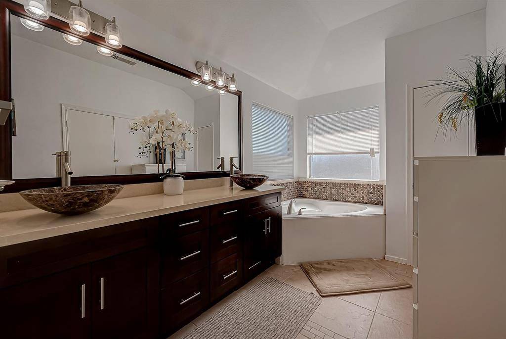 This bathroom features a spacious layout with a large mirror above a double vanity with vessel sinks. It includes a corner bathtub with mosaic tile accents, ample natural light from two windows, and modern lighting fixtures, creating a luxurious and inviting atmosphere.