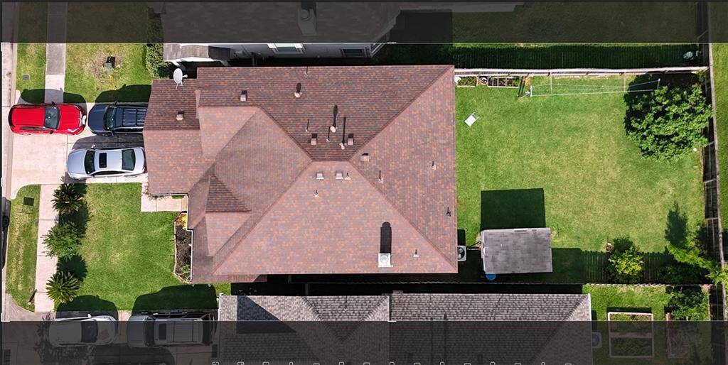 This aerial photo shows a suburban home with a well-maintained roof, a spacious backyard, and a driveway with multiple parked cars. The property is bordered by a fence and features a small structure or shed in the backyard, offering ample outdoor space for activities.