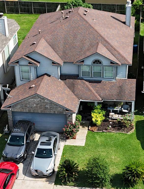 This photo shows a two-story house with a brown roof and light-colored exterior. There’s a two-car garage and a driveway with multiple cars parked. The front yard is landscaped with greenery and a small patio area. The property is fenced and appears well-maintained.