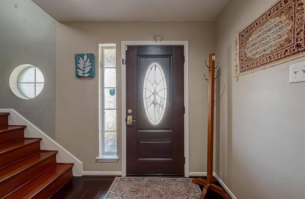 This inviting entryway features a stylish front door with decorative glass, a side window for natural light, and warm wood flooring. The neutral walls are accented with a small piece of art and a wall hanging. A wooden coat rack stands in the corner, and a carpeted staircase with a circular window adds character.