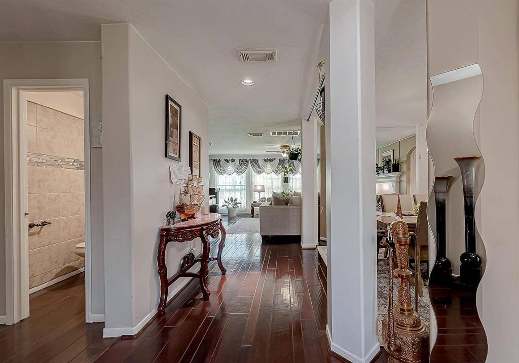 This inviting home features a welcoming hallway with polished wood floors, leading to a bright living room. A decorative console table and elegant wall mirror add charm, while a glimpse of a tiled bathroom is visible on the left. The space is well-lit and tastefully decorated, offering a cozy yet stylish ambiance.