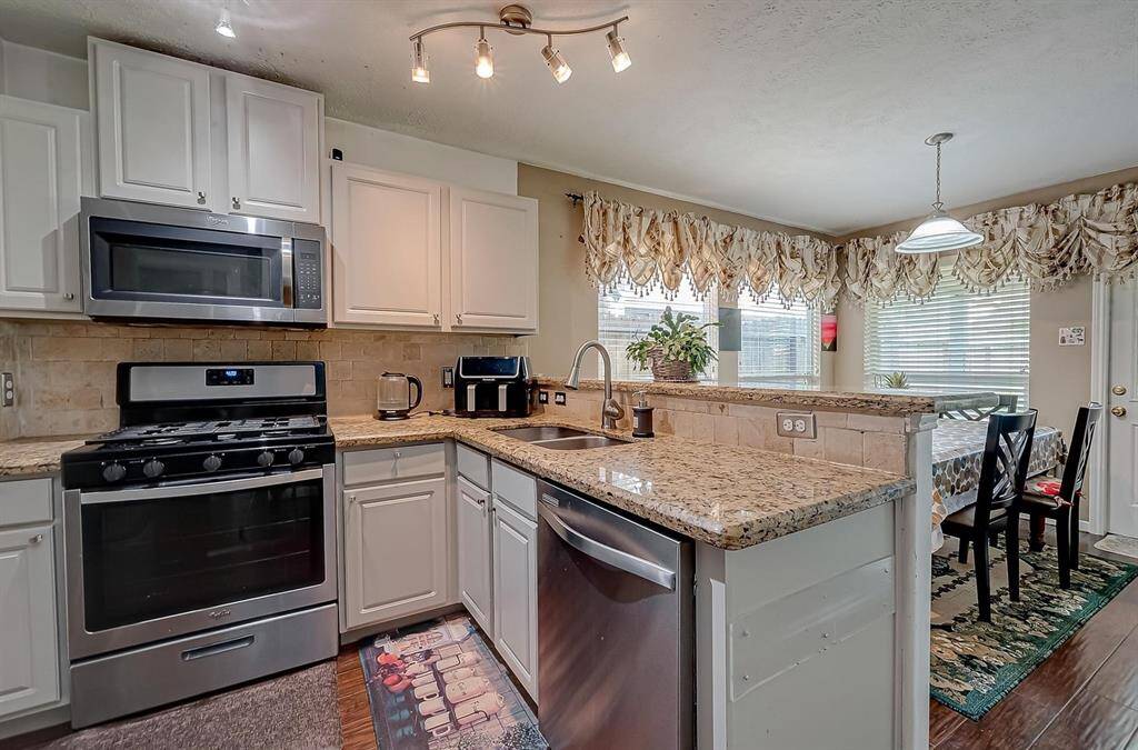 This kitchen features modern stainless steel appliances, granite countertops, and white cabinetry. It opens to a bright dining area with large windows and decorative curtains, creating a welcoming and functional space.