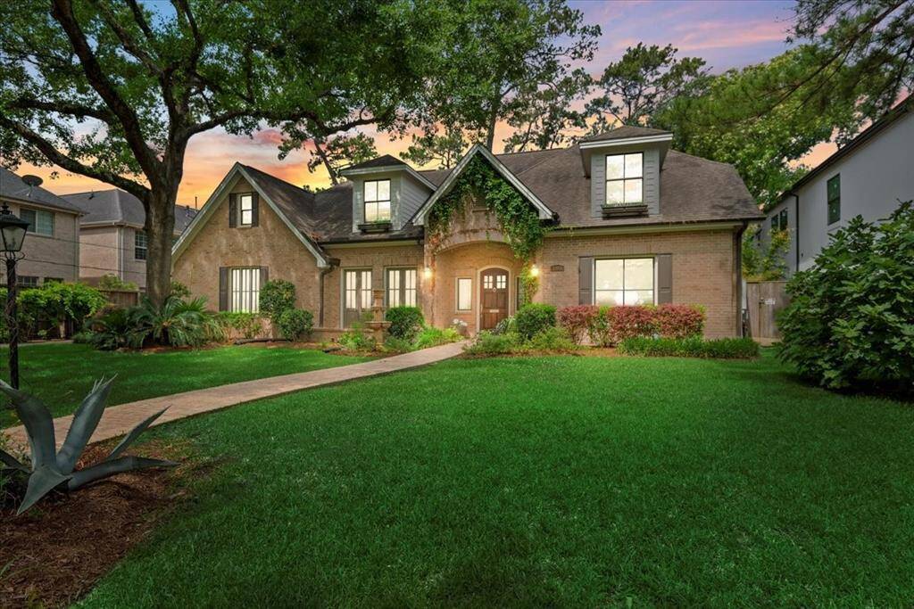 Charming two-story home with a brick exterior, nestled among mature trees. Features a well-manicured lawn, inviting front path, and elegant dormer windows.