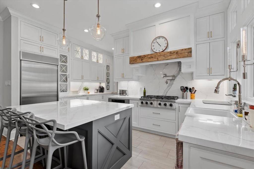 This kitchen features a modern design with white cabinetry, a large island with seating, and professional stainless steel appliances. It includes pendant lighting, a farmhouse sink, built in refrigerator, and a sleek gas range with a marble backsplash. Ideal for both cooking and entertaining.