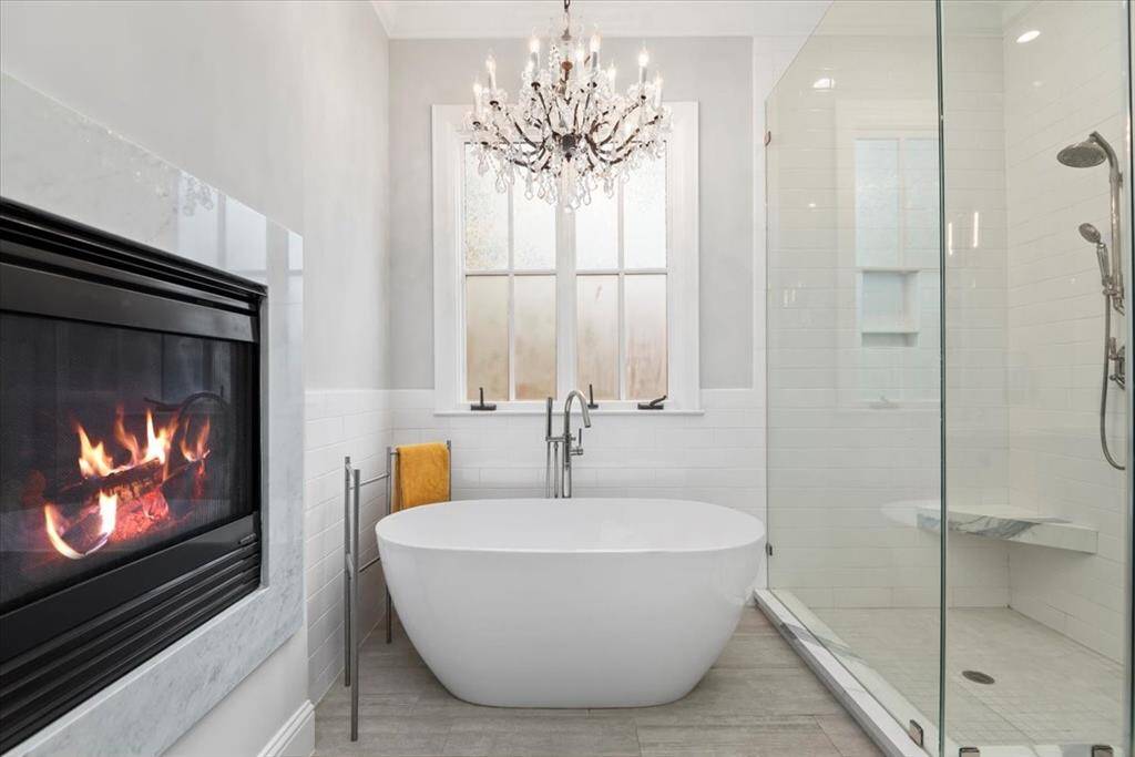This luxurious bathroom features a freestanding tub, a glass-enclosed shower, a cozy fireplace, and an elegant chandelier, creating a spa-like atmosphere.