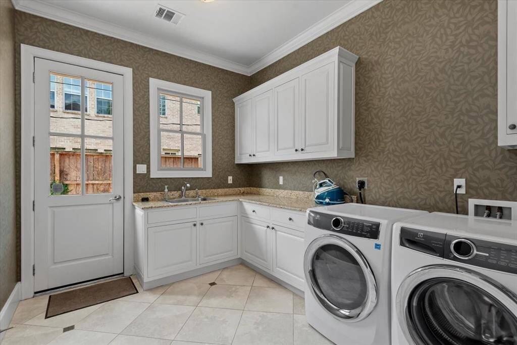 Cleverly placed next to the garage entrance, is a spacious laundry room featuring ample white cabinetry for storage, and a granite countertop with a large sink. The room is well-lit with natural light from a window and a glass-paneled door that leads outside.