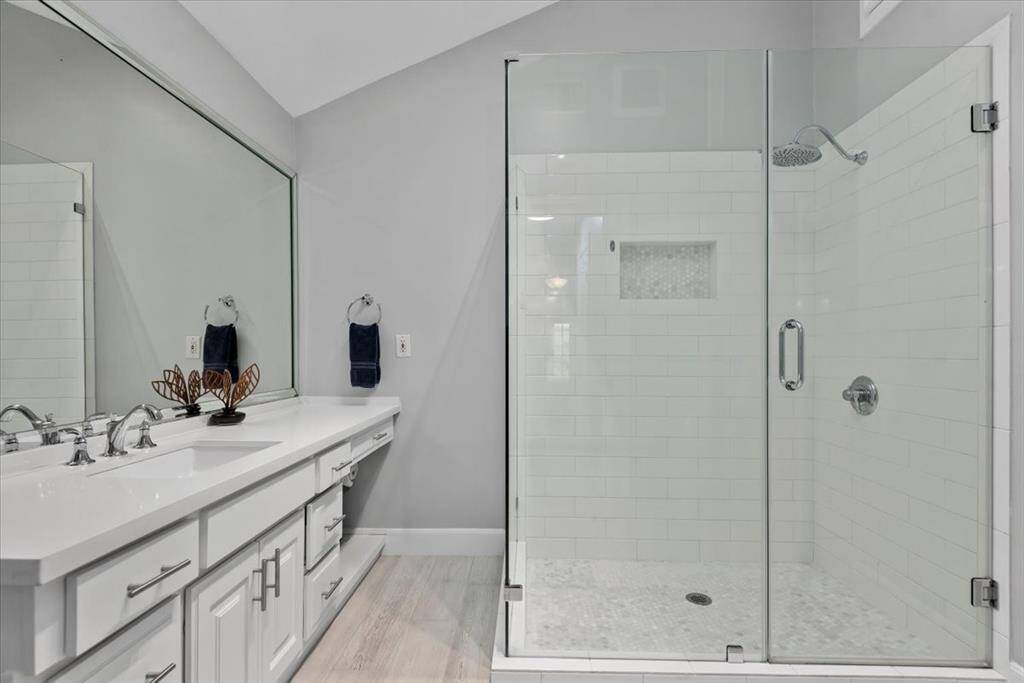 This modern bathroom features a spacious glass-enclosed shower with sleek fixtures, a large vanity with dual sinks, ample quarts countertop space, and contemporary cabinetry.