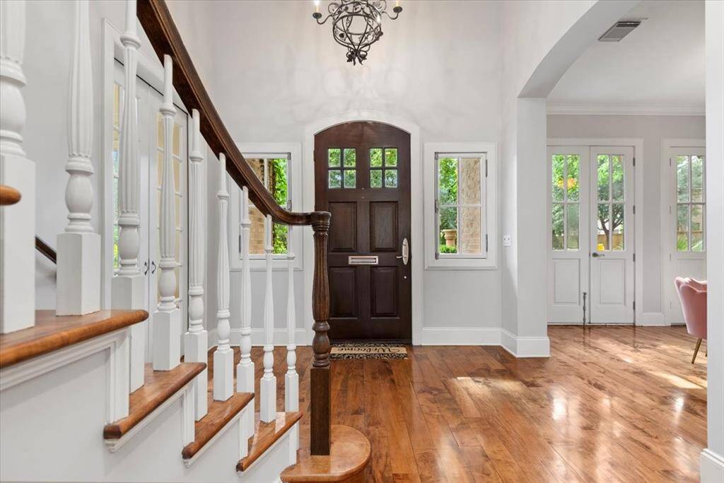 This photo showcases a welcoming entryway with rich hardwood floors, a stylish dark wood front door, and elegant white spindle staircase. Abundant natural light comes from the surrounding windows, creating a bright and inviting atmosphere.