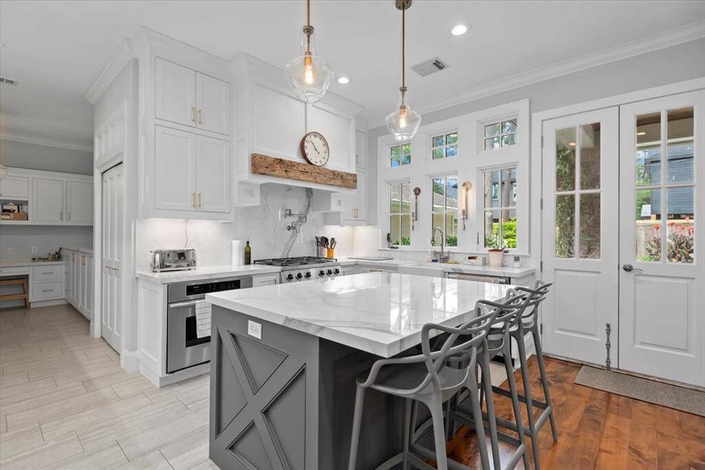 This kitchen is modern and elegant, featuring white cabinetry, marble countertops, and stainless steel appliances. It includes a central island with seating, pendant lighting, and a farmhouse sink. The space combines functionality with style, perfect for cooking and entertaining.