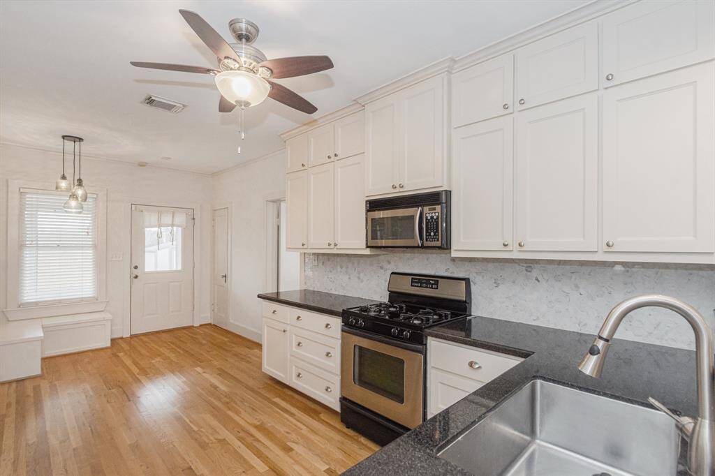 This bright kitchen features elegant white cabinetry, sleek black countertops, and stainless-steel appliances. The space is complemented by hardwood floors, a ceiling fan, and modern pendant lighting near a cozy breakfast nook by the window.