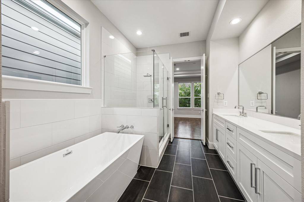 Enjoy luxurious spa-like amenities in this spacious bathroom featuring a sleek soaking tub, a glass-enclosed shower, and dual vanity with elegant finishes. Natural light filters through the large window, illuminating the pristine design and creating a serene ambiance perfect for relaxation.