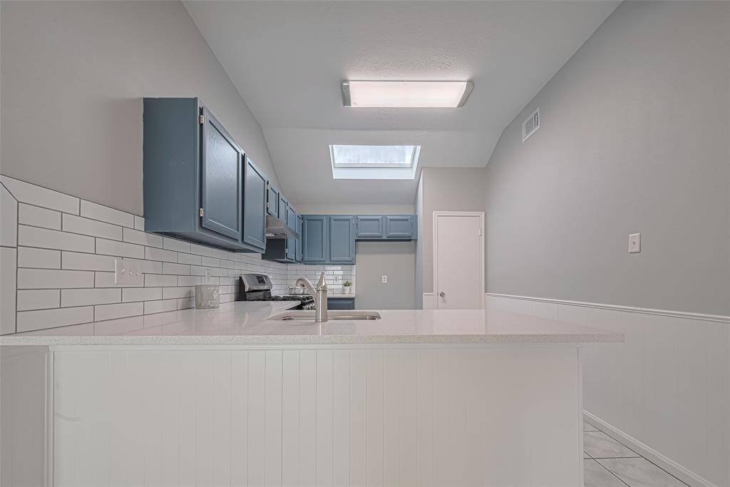 The kitchen is both calming and stylish, with blue-gray cabinetry, bright white counters and backsplash, and a skylight that pours in daylight. Clean lines and contemporary lighting give the space a polished yet cozy character.