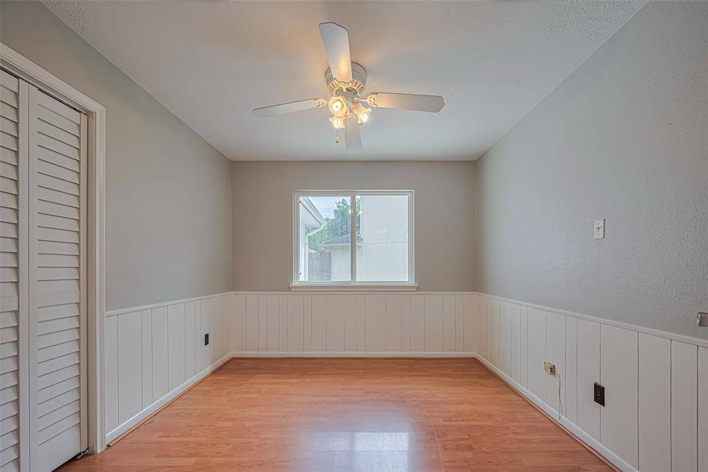 This bedroom balances warmth and clarity with sunlit wood-like floors, a paneled lower wall detail, and a generous window. A ceiling fan adds a practical touch.