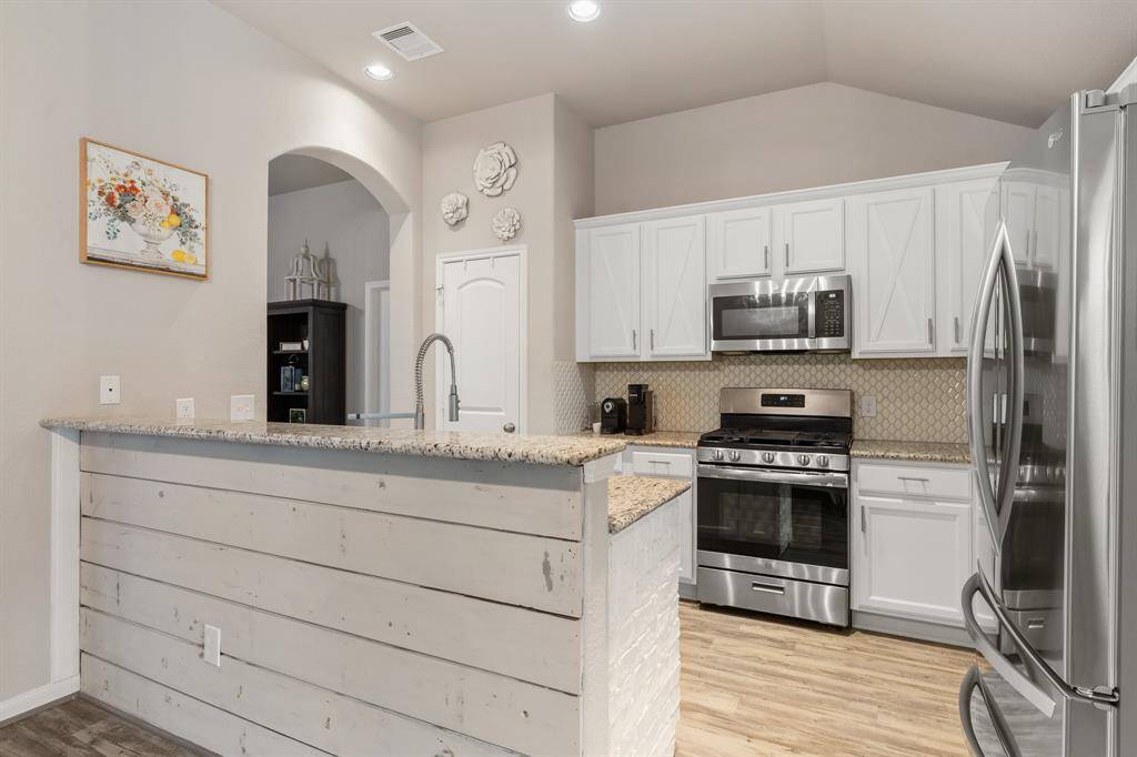This kitchen features a modern gas range, stylish white cabinetry, and granite countertops. The space is accented by a decorative backsplash and a quaint bar area with light wood paneling.