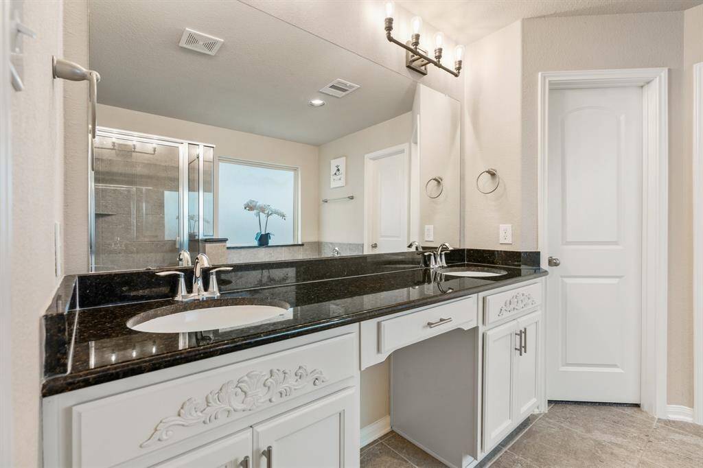 This bathroom features a dual-sink vanity with elegant black countertops and white cabinetry. It has a large mirror, modern lighting, and a spacious walk-in shower with glass doors, creating a bright and stylish space.