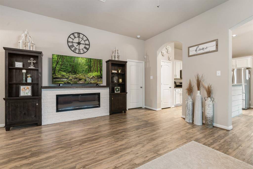 This inviting living room features electric fireplace with a sleek white brick surround. The open layout leads to a bright kitchen area.