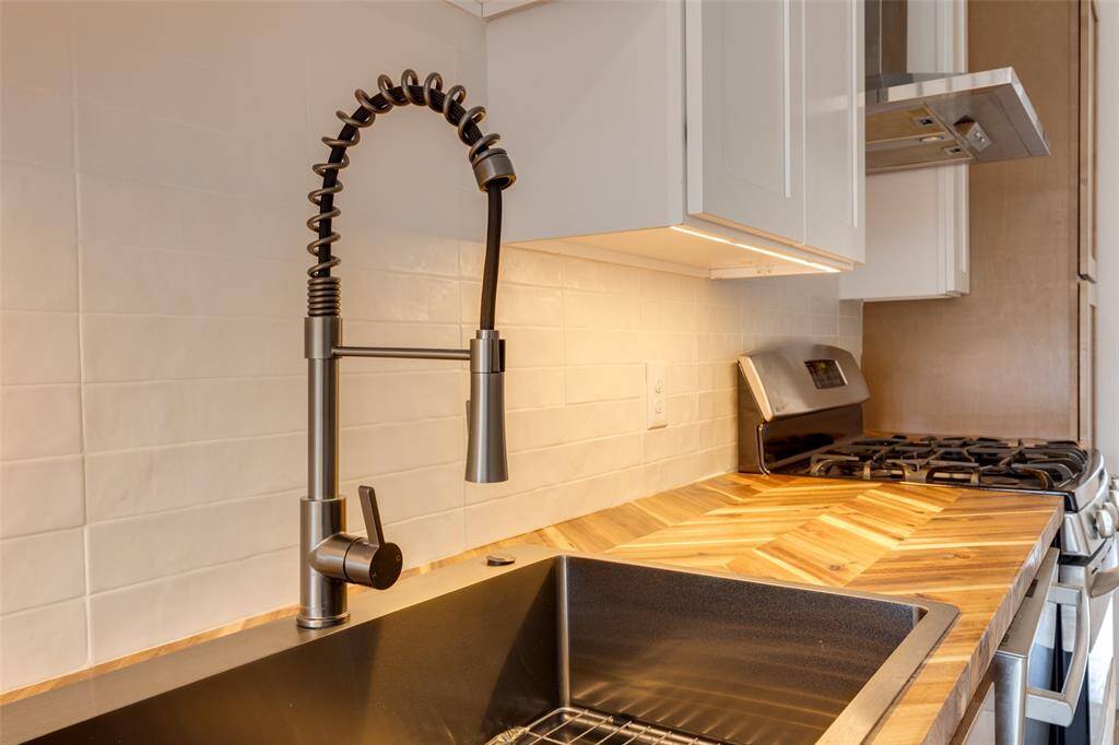 This new kitchen features a modern faucet with a flexible spout, a deep sink, and a stylish wooden countertop. The backsplash is tiled, and there is a stainless-steel 5 burner gas range with an overhead stainless-steel range hood.