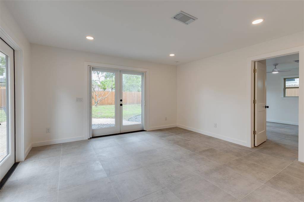 This dining room is a spacious and versatile space featuring large format tile flooring, neutral walls, and ample natural light from double glass doors leading to a fenced backyard. It connects to the adjacent primary bedroom.