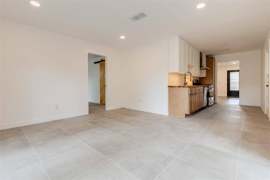 This spacious room features a modern open-plan design with neutral-colored tile flooring and recessed lighting. It includes a sleek kitchen area with wood cabinetry and stainless-steel appliances, an ideal space for a cozy dining or a second living space.
