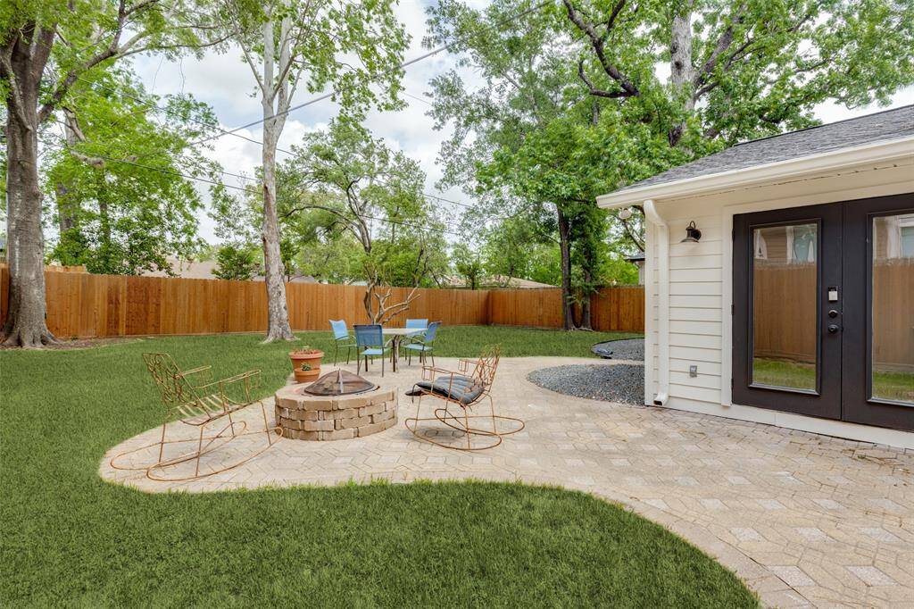 Huge backyard with a spacious paver patio featuring a central fire pit, surrounded by cozy seating area. The area is enclosed by a wooden fence and shaded by mature trees, offering a serene and private outdoor space ideal for relaxation or entertaining.