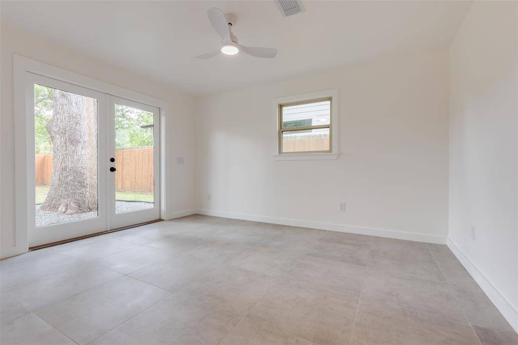 Bright primary bedroom with large format tile flooring, a ceiling fan, and French doors leading to the backyard. A small window adds extra light. Neutral colors create a modern, airy feel.