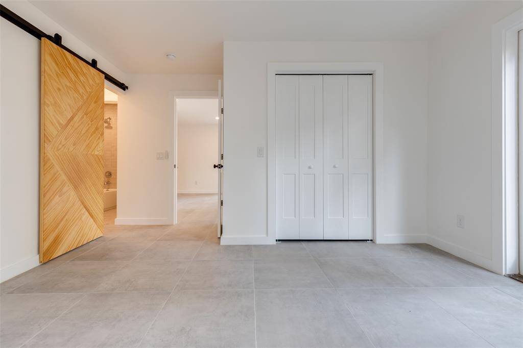 The primary bedroom features modern tile flooring, a unique wooden sliding en-suite bathroom barn door, and a double-door closet. The neutral walls and ample natural light create a bright, open space, perfect for versatile use.