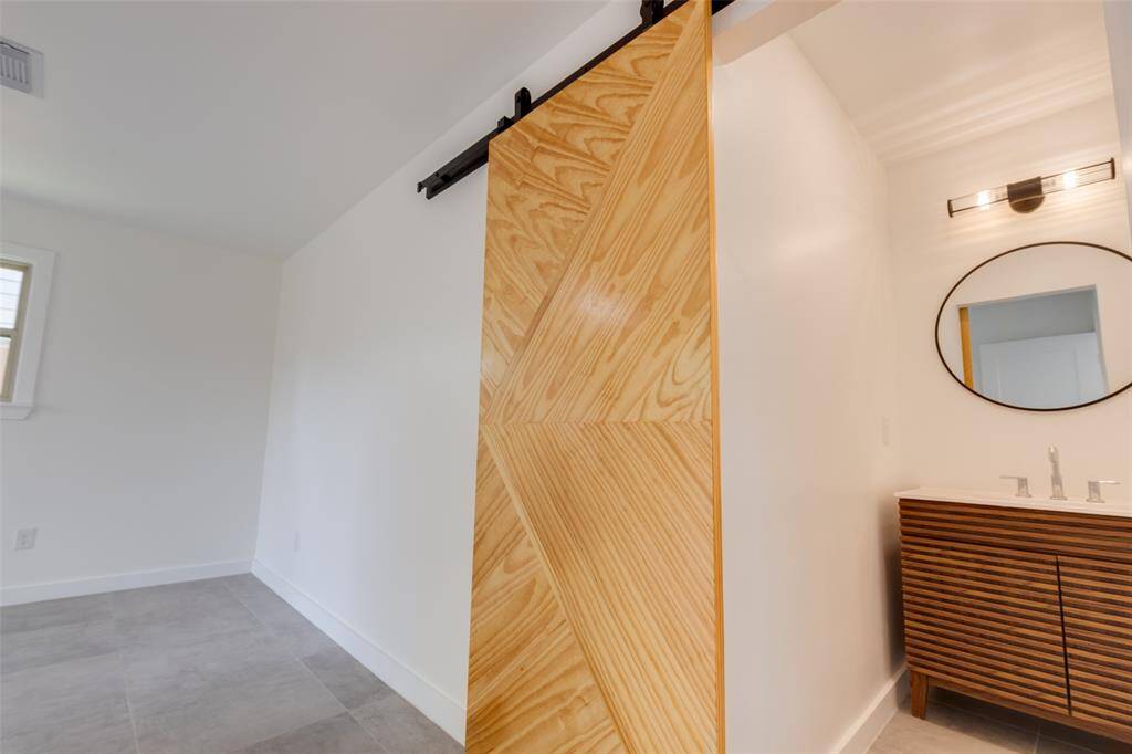 This photo features a new modern ensuite bathroom with a stylish wooden sliding barn door. Inside, there's a sleek vanity with a round mirror and contemporary light fixture, set against neutral-colored walls and flooring.