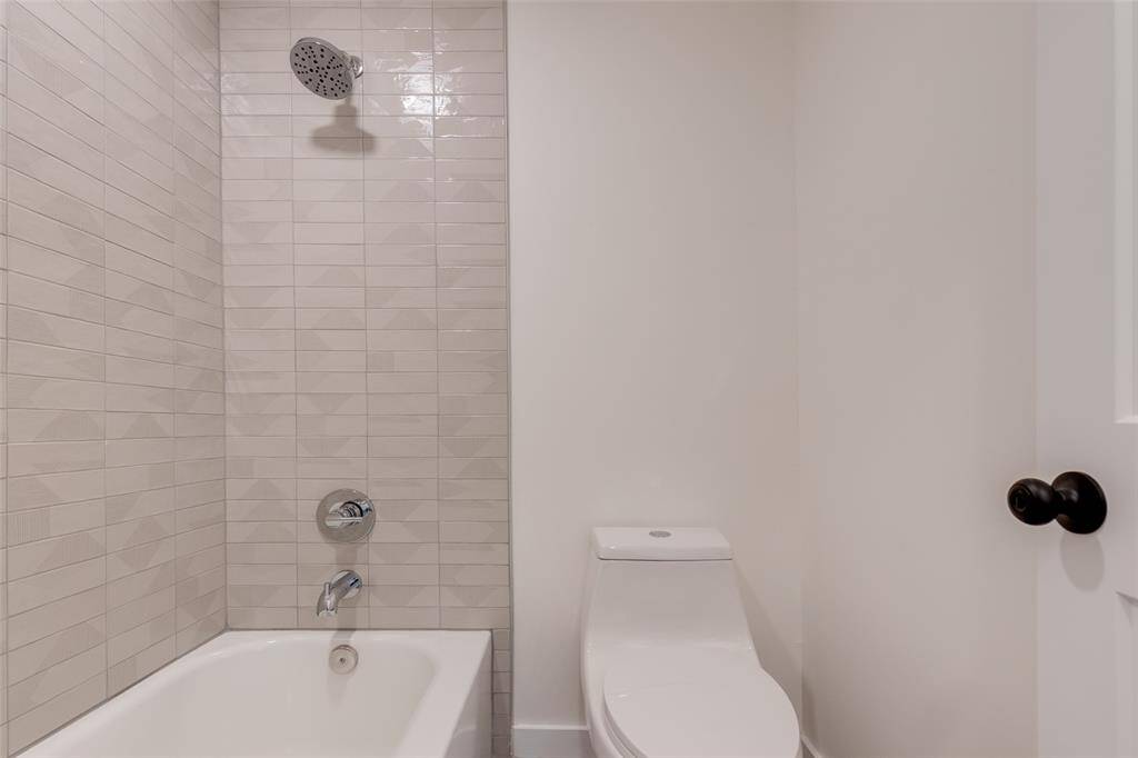 This primary en-suite bathroom features a modern design with a sleek, white bathtub and a tiled shower area. The walls are light-colored, creating a clean and bright atmosphere. A contemporary toilet complements the minimalist aesthetic.