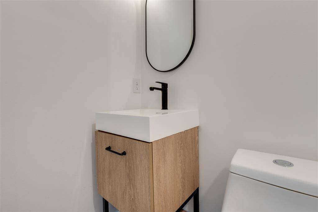 This hall bathroom features a modern vanity with a wood finish, a sleek white sink, a black faucet, and a curved mirror. The space is complemented by a minimalist design and a white toilet.