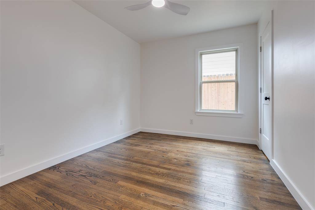 Another view of the secondary bedroom featuring fresh white walls, a window allowing natural light, and rich hardwood floors. It also includes a modern ceiling fan and a door, offering a simple and clean space ideal for a bedroom or office.