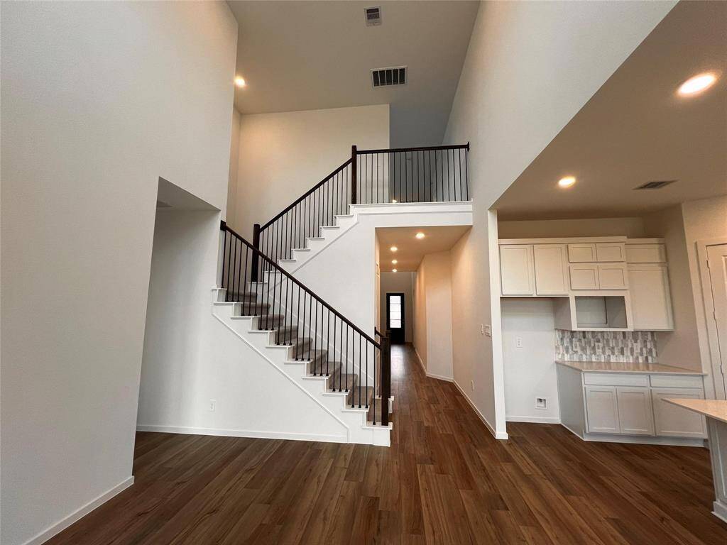 A bright and spacious living room with a high ceiling, featuring an open-concept design that flows into the kitchen area. It boasts warm wood flooring and recessed lighting, creating a welcoming and modern atmosphere
