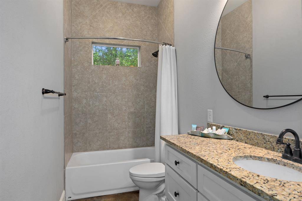 This Guest House bathroom features a sleek design with a granite countertop, a round mirror, and a tiled shower with a window for natural light. The white cabinetry and neutral color palette create a clean and modern feel.