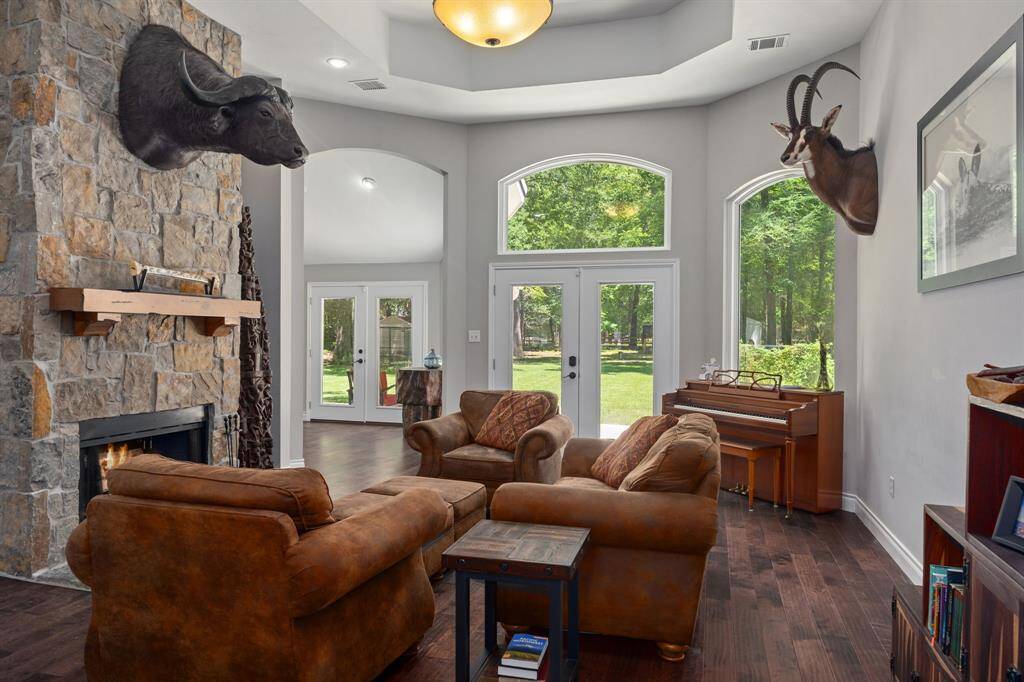 Spacious living room with large windows offering a view of a lush backyard. Features a stone fireplace with wood mantle & wood burning fireplace. Bright and inviting space for relaxation or entertaining.